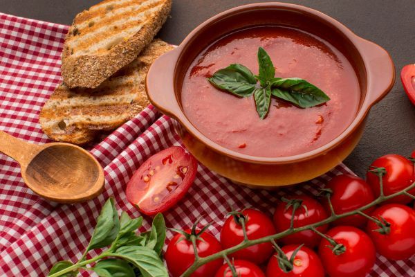 Imagem de um pote de sopa de tomate, com alguns tomates inteiros e torradas ao redor.